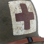 Nahaufnahme des vorderen roten Kreuzes auf dem abgenutzten weissen Feld des US M1 Combat-Medic-Helms, Lederkinnriemen des Innenhelms