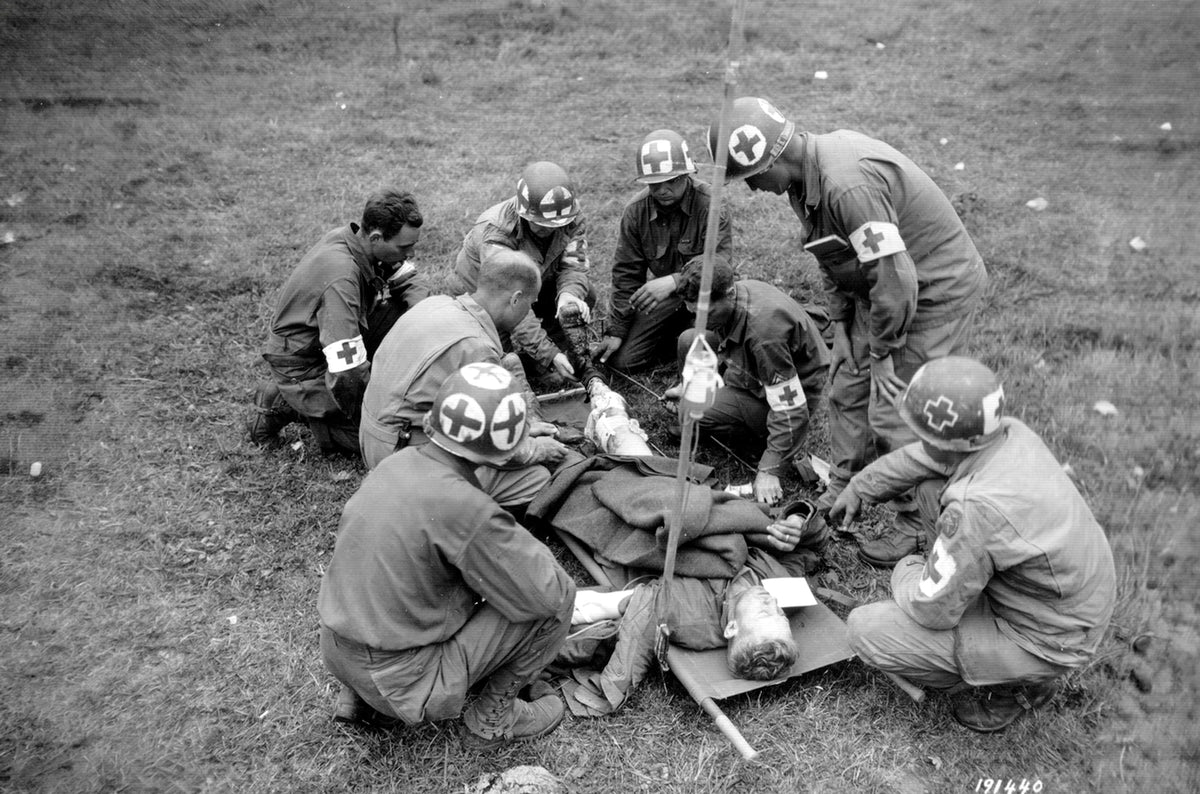 Photo d'époque de combat medics américains soignant un blessé sur le terrain, casques marqués de croix rouges et brassards sanitaires